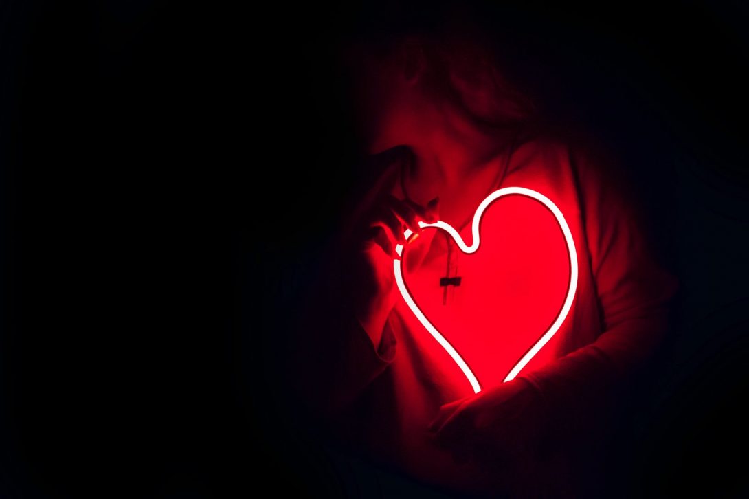 glowing red neon heart being held up to someone's chest in the dark