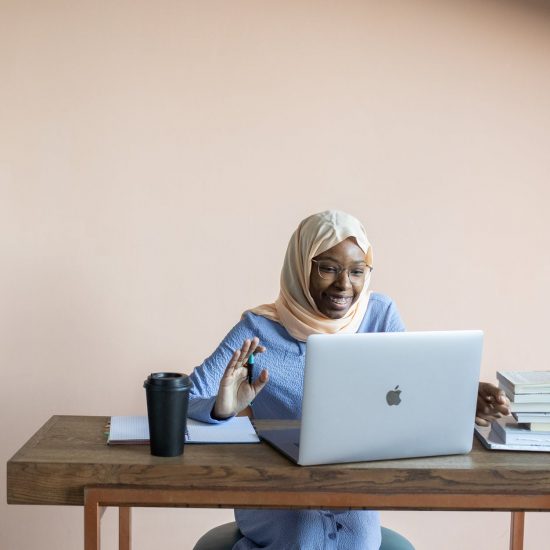Black woman wearing a hijab sitting at a wooden desk and smiling at an open laptop