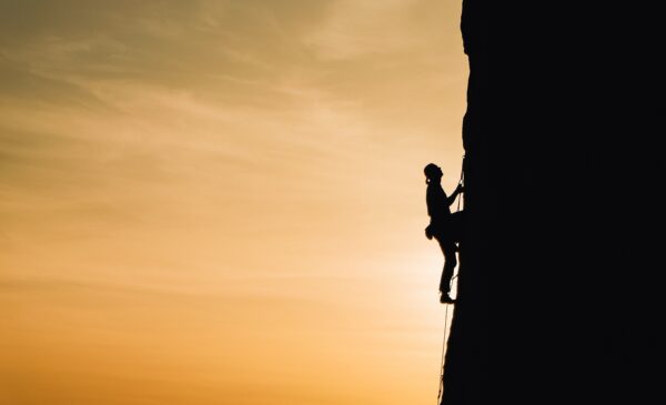silhouette of a person climbing up the side of a  mountain while the sun sets behind them